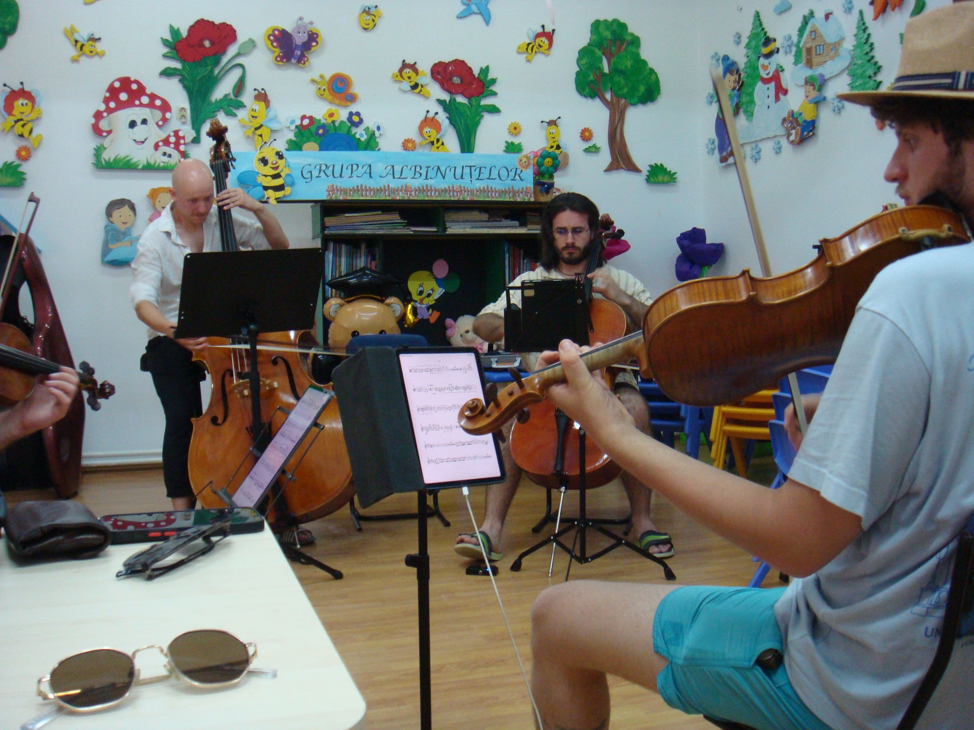 Three musicians rehearsing in a kindergarten classroom, a wall of cartoon bees and flowers behind them, a violin scroll in the foreground.